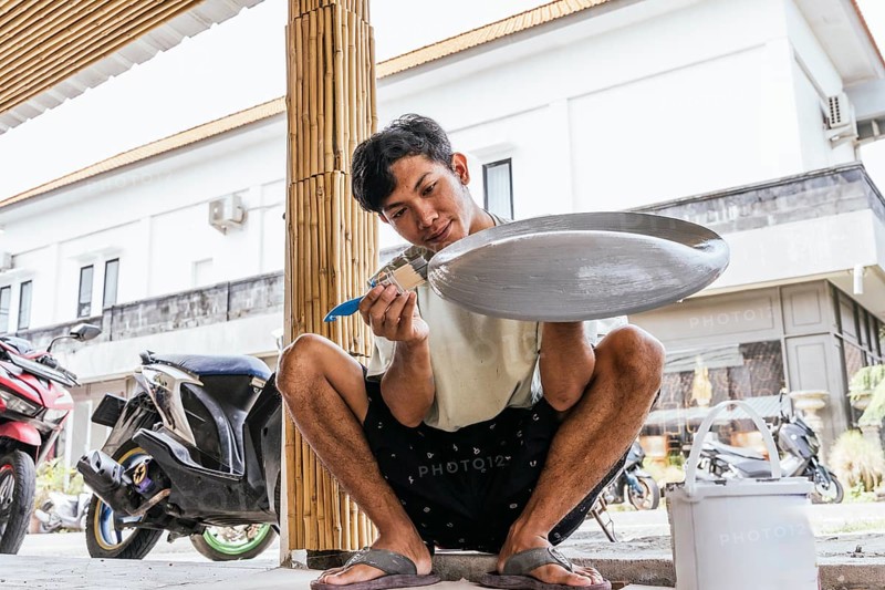 A Balinese artist skillfully paints a sculpture outdoors, surrounded by local architecture and bamboo elements. The scene highlights cultural artistry and craftsmanship