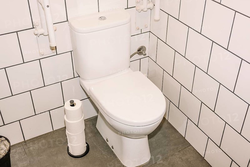 A clean white toilet in a coffee shop restroom, surrounded by white subway tiles and a gray floor. A stack of toilet paper rolls is placed nearby, ensuring hygiene and convenience for customers