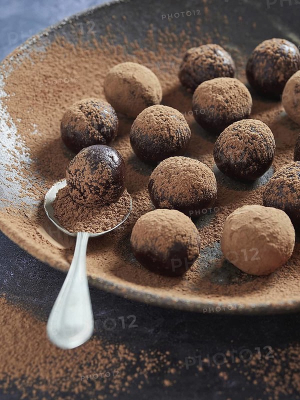 Chocolate bliss balls dusted with cacao on a dark plate with a spoon. Healthy homemade treat, sugar free truffles made of dates, cocoa and nuts. Energy balls. Step by step cooking. Recipe