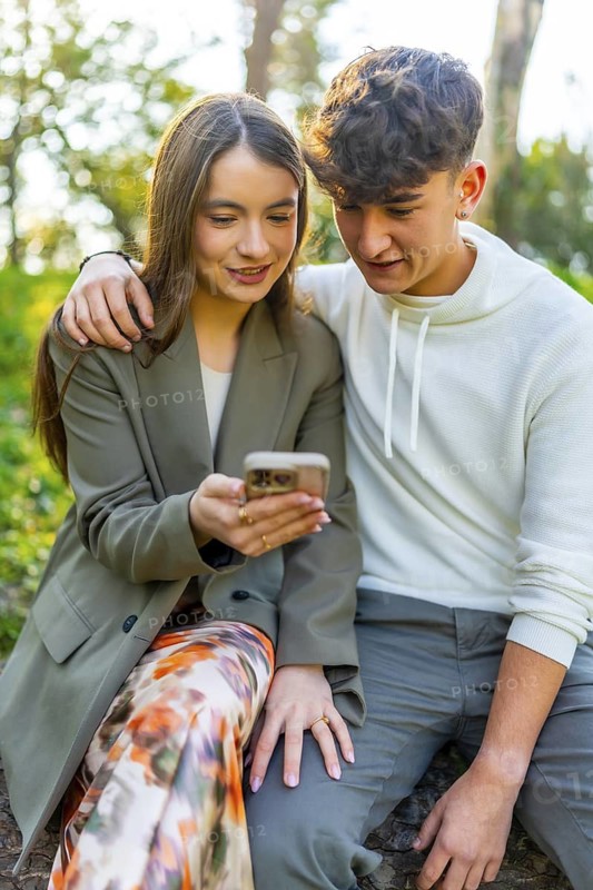 Smiling young couple sitting in a park, sharing a moment while looking at a smartphone, surrounded by lush greenery and sunlight