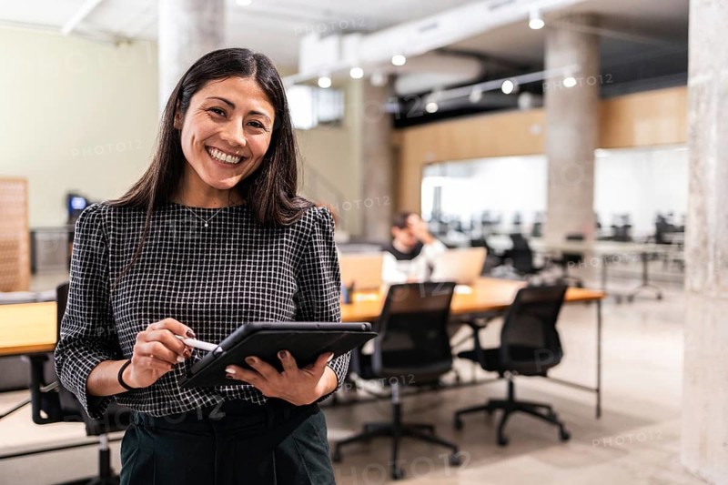 A smiling woman in a professional setting holds a tablet, embodying teamwork and productivity in a modern office environment Collaborators work in the background