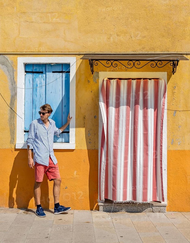Young man leaning against house wall, yellow house facade with entrance door with red and white curtain and windows with blue shutters, colourful houses on the island of Burano, Venice, Veneto, Italy