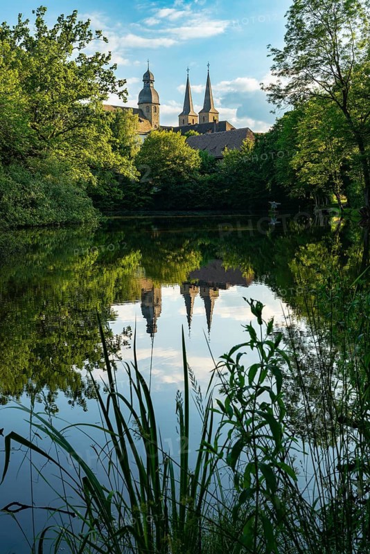The towers of the monastery church of Marienmünster Abbey are reflected in a tranquil lake surrounded by trees, North Rhine-Westphalia, Germany