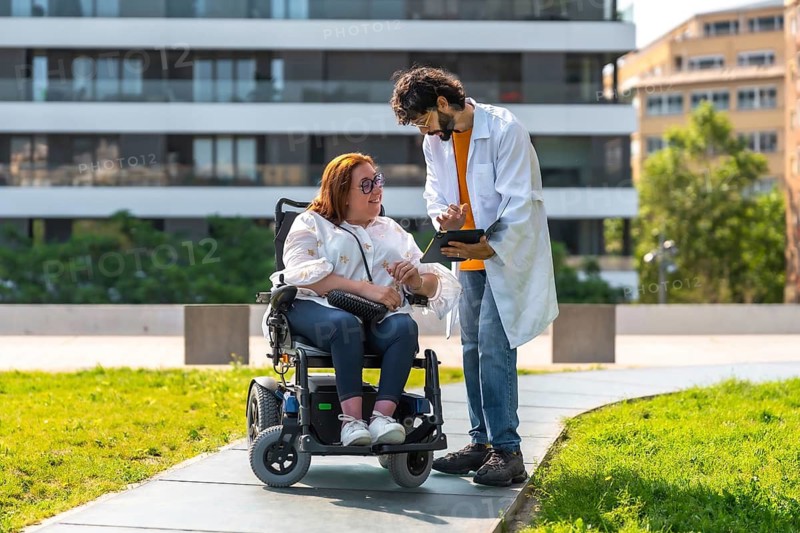 Doctor holding tablet explaining diagnosis to smiling woman sitting in wheelchair in urban park