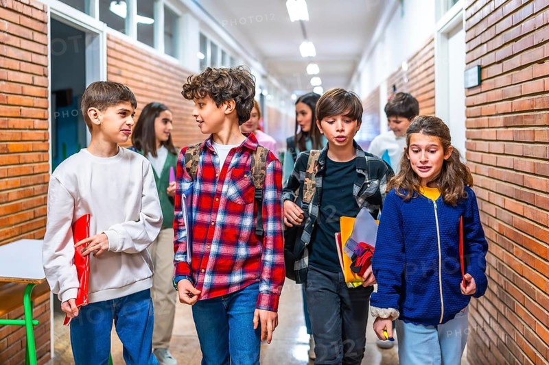 Elementary school students carrying backpacks and notebooks walking down school hallway between classes, chatting and smiling
