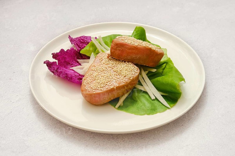 Two pieces of sesame-coated tuna fish are on a plate with a mix of vibrant greens and thinly sliced vegetables. The dish showcases a fresh and inviting presentation