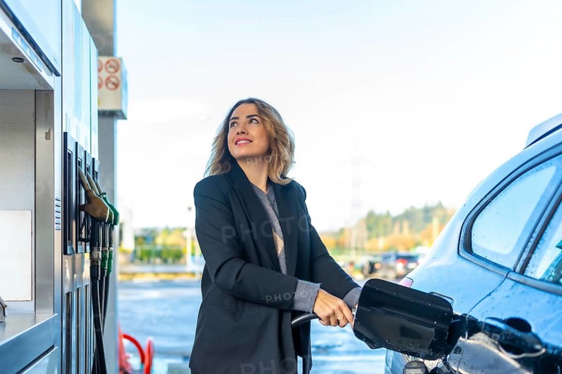 Woman standing at a gas station, feeling happy and looking up while filling car with fuel, representing independence, travel, and modern automotive transportation