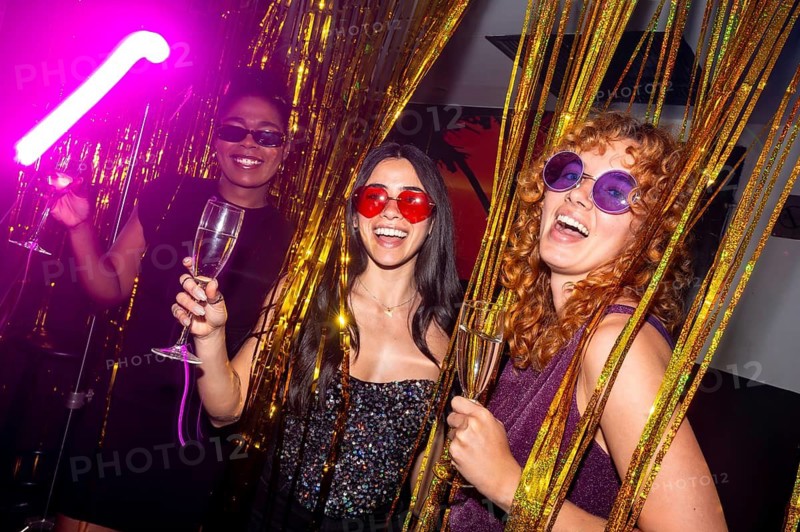 Three cheerful young women with colorful sunglasses are holding glasses of sparkling wine, enjoying a vibrant disco party atmosphere with golden tinsel decorations and neon lights