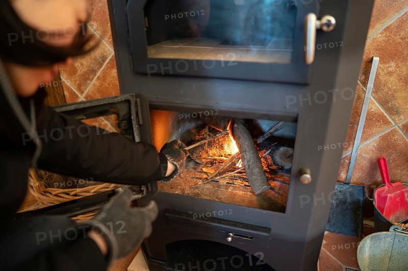 Cropped image of an Asian person wearing gloves as they stoke a wood burning stove, ensuring the flames wrap around the logs efficiently