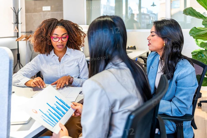 Three businesswomen are discussing over financial charts sitting at a desk in a modern coworking office
