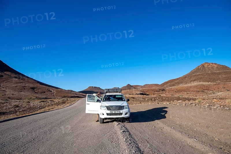 Tourist with off-road vehicle on gravel track leads through barren desert landscape, sandstone rock formation, silhouettes of mountains against the light, Damaraland, Kunene, Namibia
