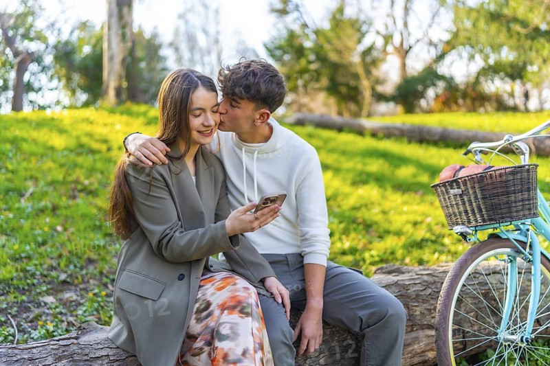 Happy couple sitting on a log in a park, sharing a moment with a smartphone, bicycle nearby, surrounded by lush green nature