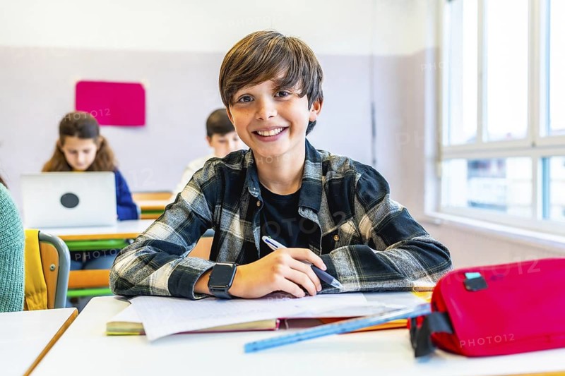 Happy student smiling while sitting at desk in classroom during lesson, with classmates in background using laptops