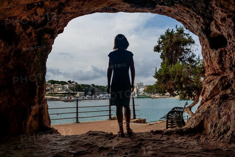 A woman stands in a cave looking out at the water. The cave is dark and the woman is silhouetted against the light. Scene is peaceful and contemplative