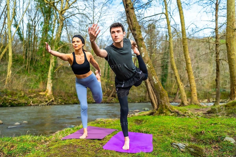 Man and woman practicing yoga in nature, performing the natarajasana pose on mats near a river, surrounded by trees