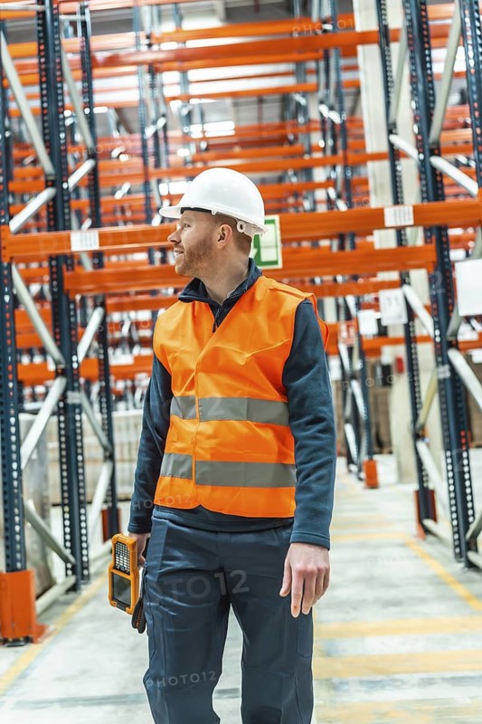 Warehouse worker wearing high visibility vest and hardhat walking through logistics center while holding handheld scanner