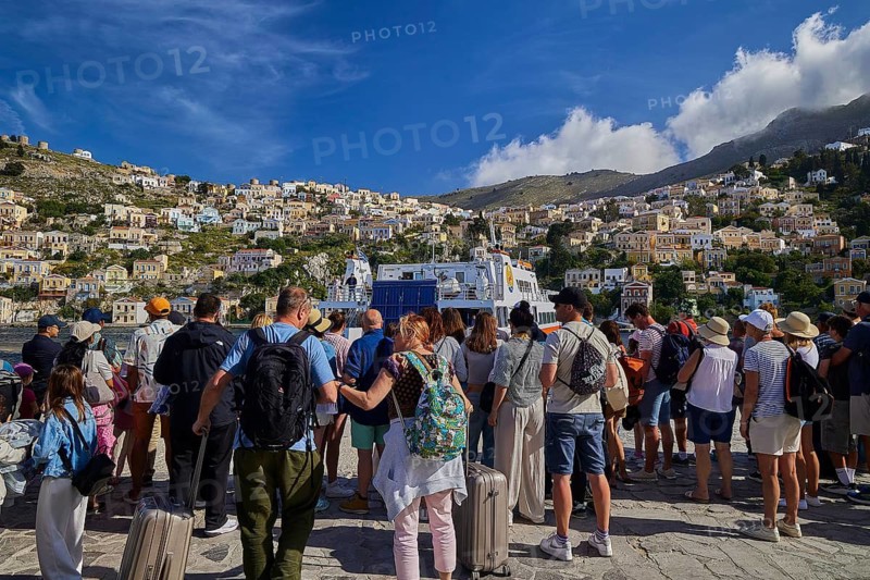 People waiting in a coastal town with colourful houses on a hill under a blue sky with clouds, Symi Town, Symi, Dodecanese, Greek Islands, Greece
