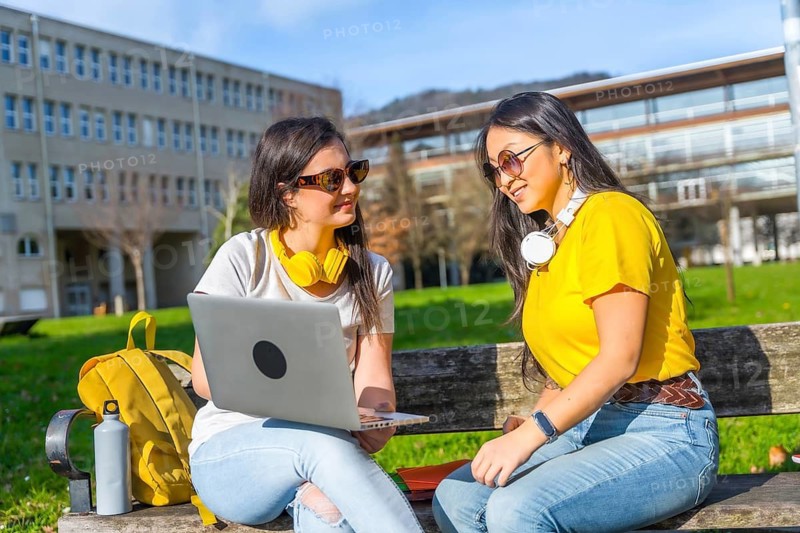 Two young women studying using laptop and headphones sitting outside the university