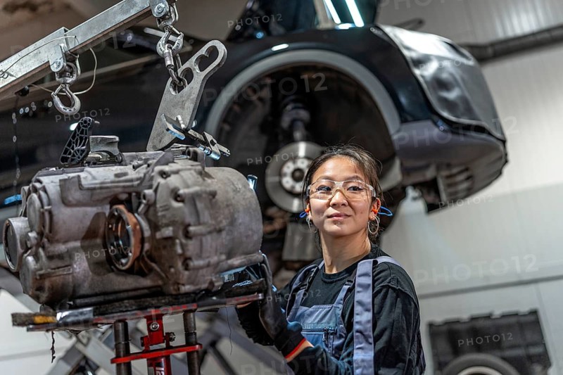 Young woman working on a gearbox with a crane in a professional car service, showcasing skills in automotive repair and maintenance
