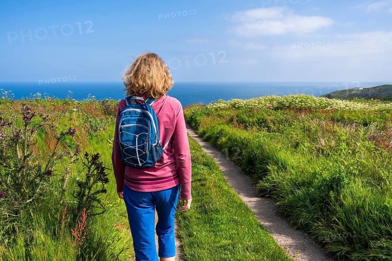 A hiker with a rucksack on a coastal path in Cornwall. View of the sea. Wildflowers in bloom. Near Gurnard's Head, Cornwall, England, Great Britain