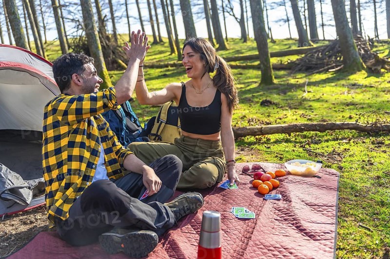 Happy couple sharing a joyful high five while camping, playing cards, and enjoying a picnic near their tent in a serene forest setting