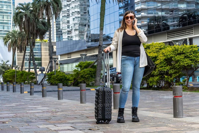 Young businesswoman pulling a suitcase while talking on her smartphone, walking confidently along a bustling city street surrounded by modern office buildings on a sunny day