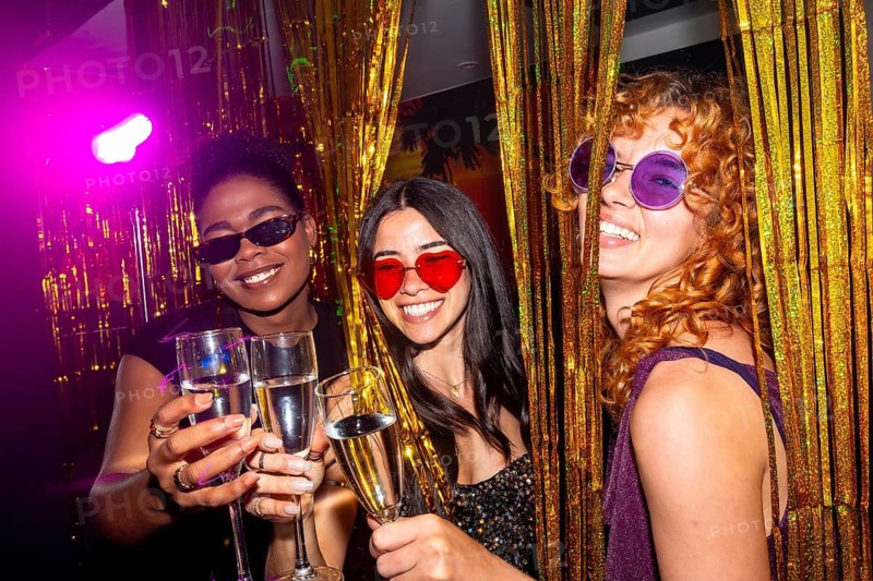 Three multi ethnic female friends are toasting with champagne flutes at a nightclub, enjoying the nightlife and wearing colorful sunglasses