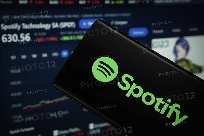 Dhaka, Bangladesh- 18 March 2025: Spotify logo is displayed on a smartphone screen, with a stock graph webpage visible on a computer screen in the background