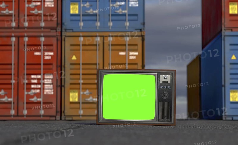 Vintage television with chroma green screen resting on shipping dock asphalt amongst stacked shipping containers