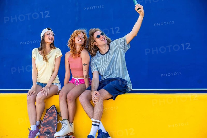 Three cheerful friends taking a selfie with a smartphone while sitting on a yellow wall with a blue background