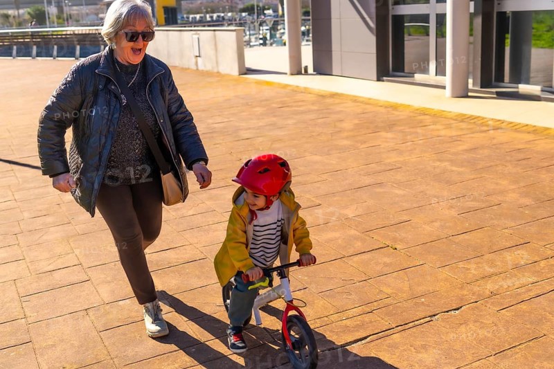 Grandmother jogging behind her grandson riding a balance bike on a paved sidewalk next to a modern building