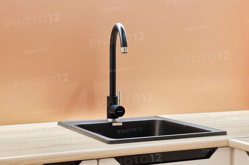 A modern kitchen sink is showcased with a sleek, high-arch faucet featuring a single handle. The sink is set in a light-colored countertop and against a warm, beige wall, emphasizing minimalism