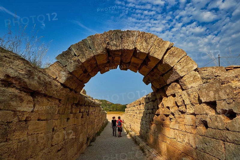 Crypt, vaulted corridor to the stadium, two people walk through an old stone arch wall under a broad blue sky, Archaeological Site, Ancient Olympia, Olympia, Peloponnese, Greece