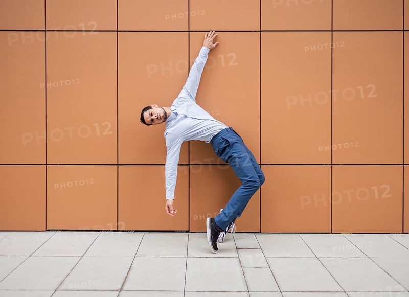 A Cuban male dancer leans artistically against a brown tiled wall, showcasing his dance skills. He is wearing a blue shirt and dark pants, capturing movement and style