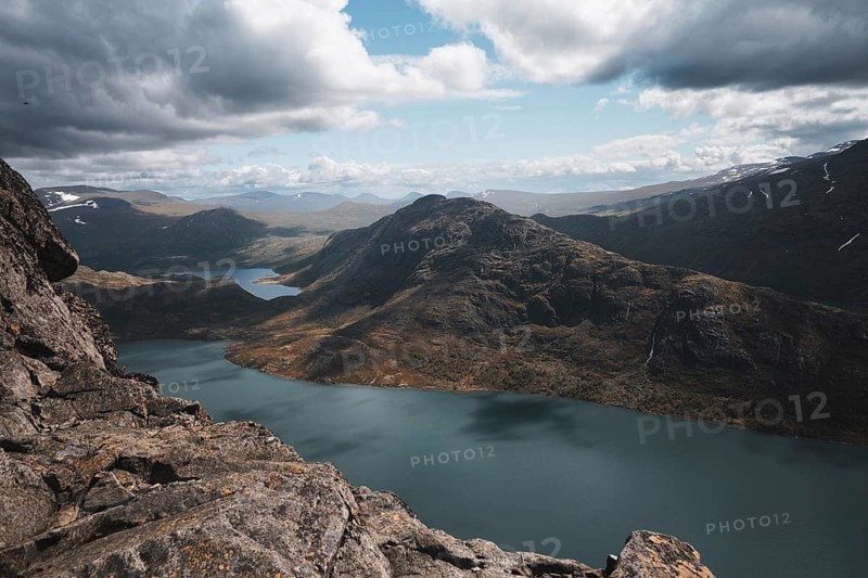An expansive view of Norway's mountainous terrain, featuring a pristine lake surrounded by rugged peaks The rocky foreground frames this dramatic and awe-inspiring Nordic landscape