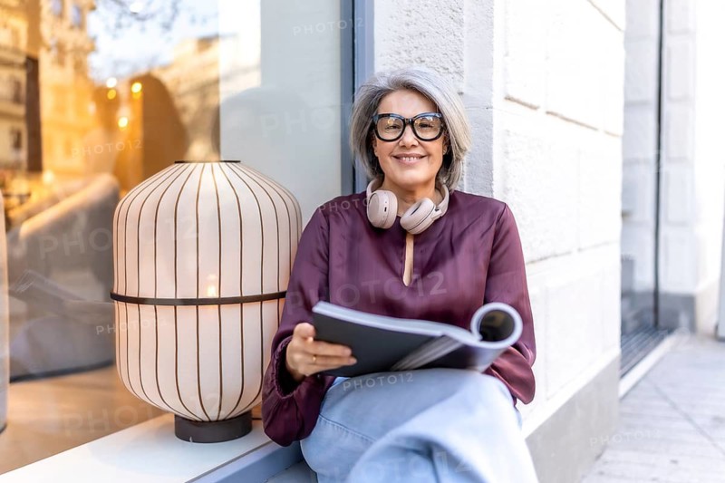 Mature woman sitting casually on a window ledge, smiling warmly at the camera while holding an open book and wearing headphones around her neck, enjoying a moment of leisure in the urban setting