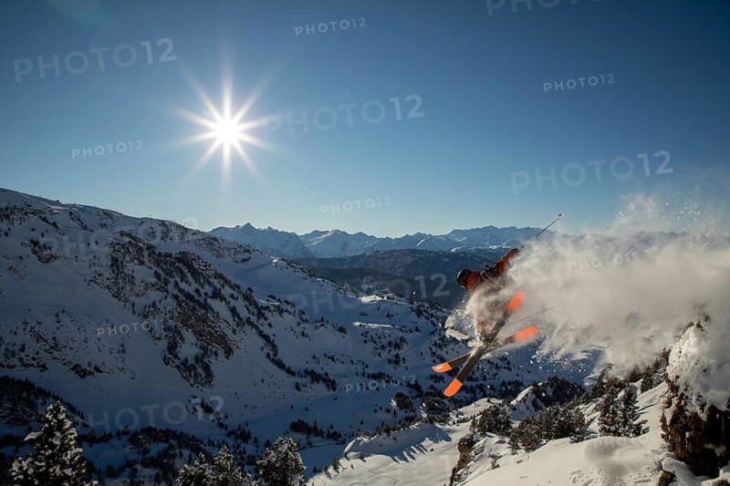 Dynamic freeride skiing scene with skier mid-jump, creating a spray of snow against a backdrop of majestic mountains and vibrant blue sky, capturing winter adventure spirit