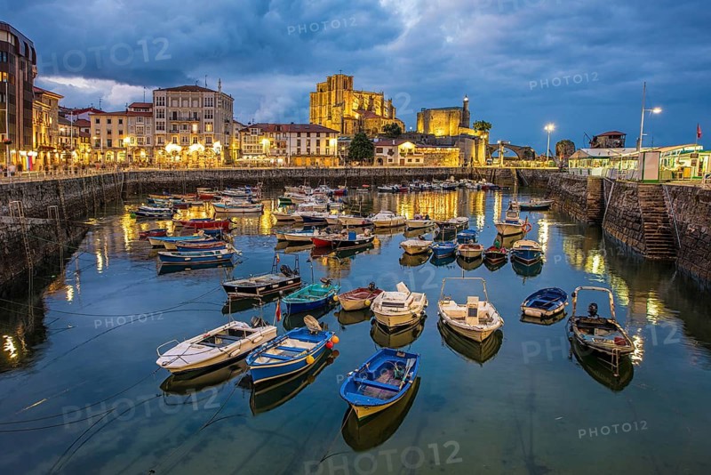 Tranquil evening view of a Castro Urdiales marina, showcasing colorful boats reflected in the calm water under an overcast sky, with illuminated historic stone buildings in the background