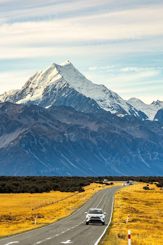 A picturesque view of Mount Cook, New Zealand, during autumn. The winding road and golden fields enhance the beauty of the snow capped peak under a serene sky