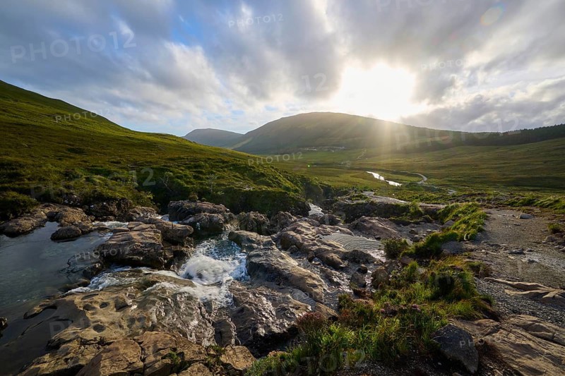 River flowing through rolling landscape in the warm light of sunset under cloudy sky, Fairy Pools, Grula, Isle of Skye, Scotland, United Kingdom, Great Britain
