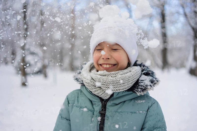 A joyful child with a white hat and scarf plays in the snow, surrounded by falling snowflakes. The wintry scene captures pure happiness and the beauty of a snowy day