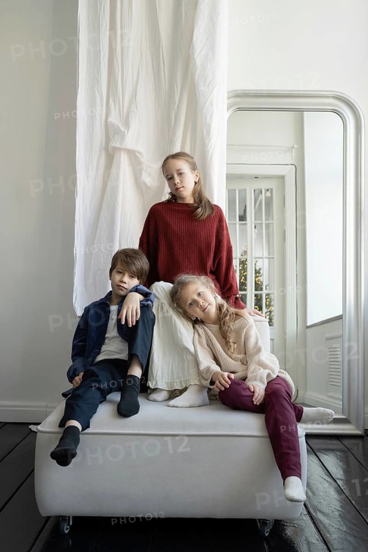 Three children dressed in casual, cozy attire pose together in a bright, minimalistic room The calm setting features soft lighting and simple decor