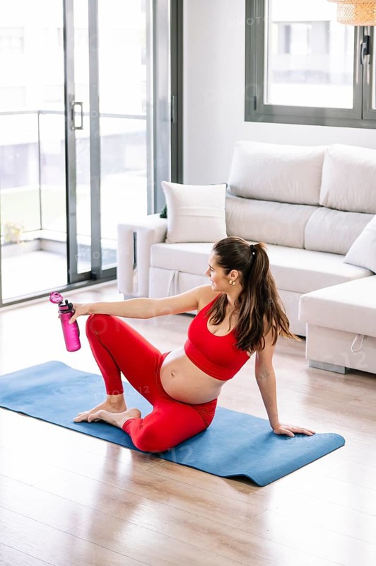 Pregnant woman practicing yoga in a sunny living room while wearing a red outfit, sitting on a blue yoga mat, holding a water bottle, promoting wellness and relaxation