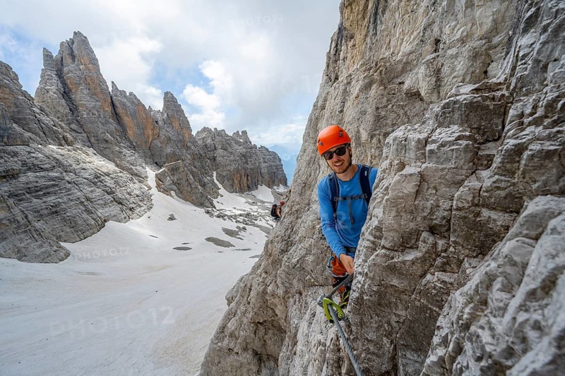 Mountaineer climbs on a steep rock face in the Via Ferrata dell'Ideale via ferrata, spectacular mountain landscape with steep rock faces, remnants of the Vedretta d'Ambiez glacier in the back, Brenta Mountains, Brenta-Adamello Nature Park, Trentino, Italy