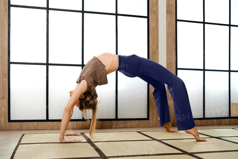 A woman is in a yoga studio practicing a backbend pose. She is using a mat. The studio has large windows that let in sunlight. The time appears to be midday, creating a bright atmosphere