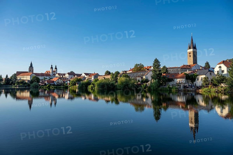 A wide panoramic view of the historic town of Telc with its colorful buildings and church towers reflected in the calm surface of a pond under a clear blue sky in the Czech Republic