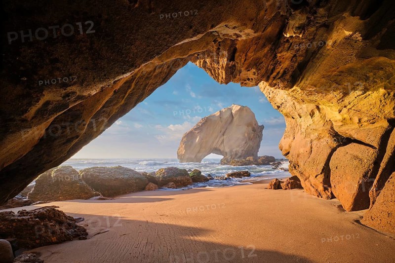 Discover the stunning Penedo do Guincho rock arch framed by a cave, with ocean waves and sandy beach at sunset in Portugal