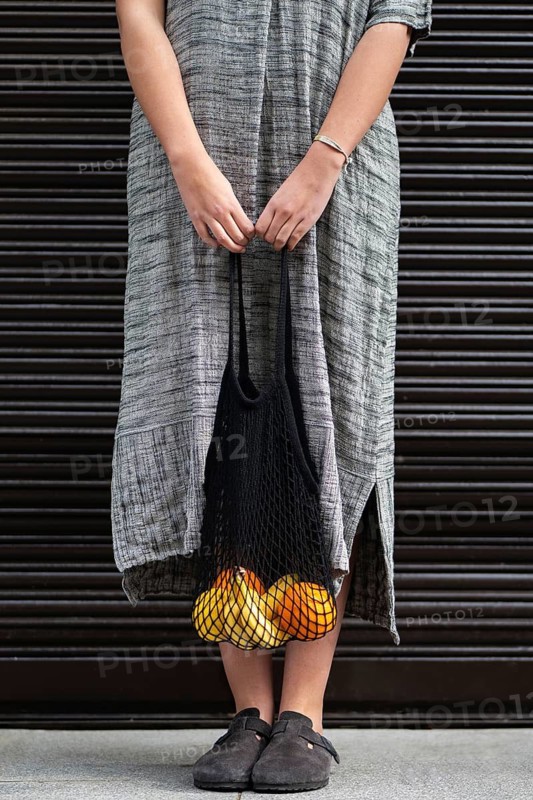 A woman in a long, gray dress carries a reusable mesh bag filled with bananas and oranges. The individual walks on a sidewalk, wearing black slip-on shoes
