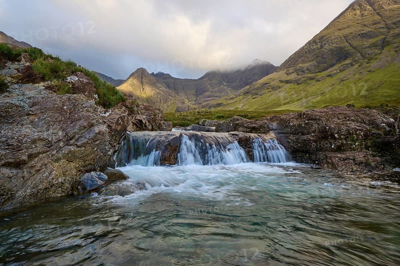 Waterfall with adjacent rocks in a green mountain landscape under cloudy sky, Fairy Pools, Grula, Isle of Skye, Scotland, United Kingdom, Great Britain