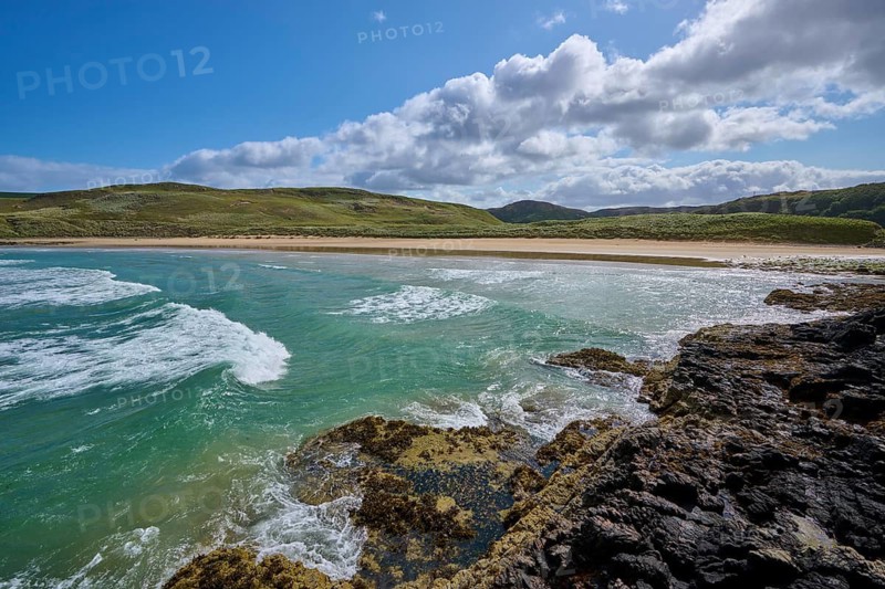 Waves crash against rocky coast against sandy beach and green hills under cloudy sky, Farr Bay, Bettyhill, Thurso, Scotland, United Kingdom, United Kingdom
